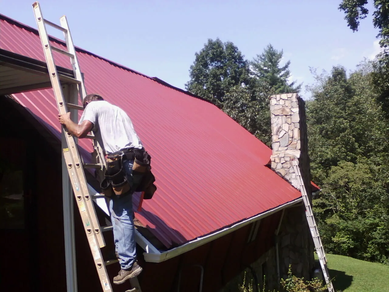 Roofer on a ladder beside a red metal roof and stone chimney — Soffit & Fascia Repair by Roofing Brothers in Lewiston